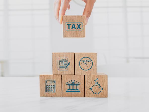 6 wooden building blocks each with a tax related image on such as the word 'TAX' a document of 'TAXES', a clock with the words 'TAX TIME', a calculator, a bank, a piggy bank. 