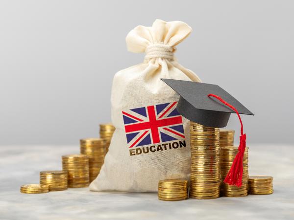 A white sack with a union jack printed on the front, the word 'EDUCATION' typed underneath in back text. next to the sack are stacks of coins, one stack is wearing a tiny graduation cap. 