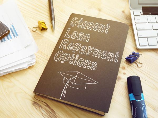 A desk with a keyboard, paperwork, various stationary and a black book with a graduation cap drawn on the front in white pen, the text on the front of the book reads 'STUDENT LOAN REPAYMENT OPTIONS'. 