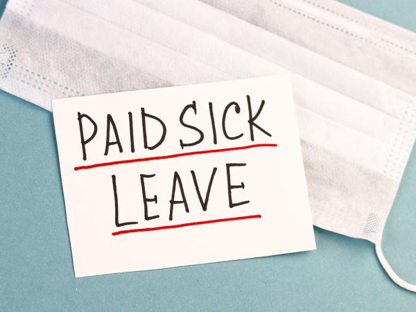a face mask and piece of white paper with the words 'PAID SICK LEAVE' written on it  against a blue background. 