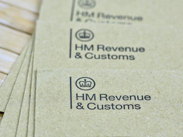 a pile of brown envelopes on a wooden table, each envelope has 'HM REVENUE & CUSTOMS' printed in the top left corner along with the HMRC logo. 