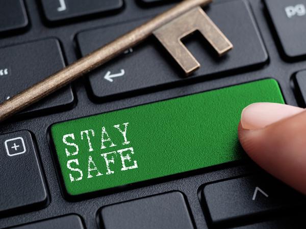 A keyboard with a Green key, a person is pressing this key. The key has white text on it that reads 'STAY SAFE' an old fashioned brass coloured key is placed on top of the keyboard. 