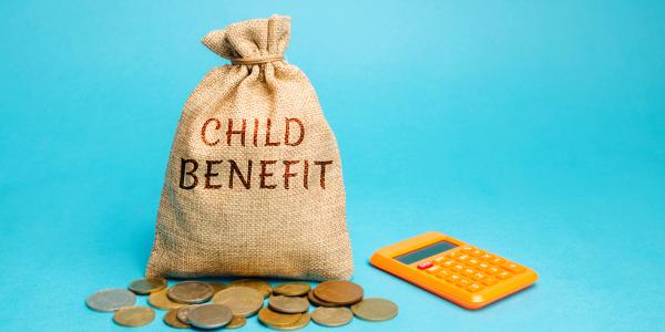 A burlap sack, an orange calculator and a scattering of coins against a blue background. On the sack the words 'CHILD BENEFIT' are written.