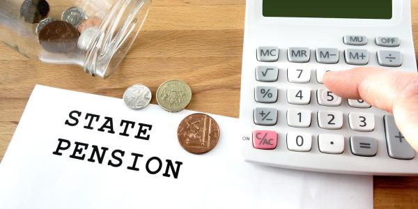 a sheet of white paper with the words 'STATE PENSION' typed on it, next to this is a glass jar of coins tipped onto its side, a few of the coins have fallen out onto the paper, a person is typing on a calculator at the right hand side. 