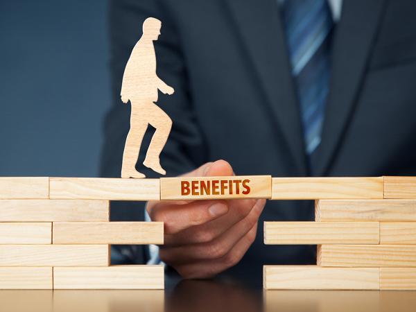 wooden blocks making a bridge with a gap in it, one block is being held in place to allow the wooden figure to cross, on this block the word 'BENEFITS' is written on the side.