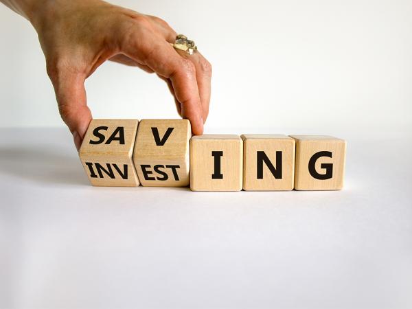 5 wooden blocks, a person is tilting the first 2 blocks so 2 sides of it can be seen, the final 3 blocks have the letters 'ING' on them, the first 2 show 'SAV' to read 'SAVING' and 'INVEST' to read 'INVESTING'