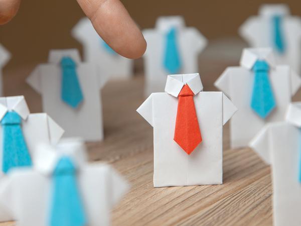 A table of origami shirts made from white paper, each wearing a little blue tie except for one that is wearing a red tie, a person is pointing to the origami shirt wearing the red tie.