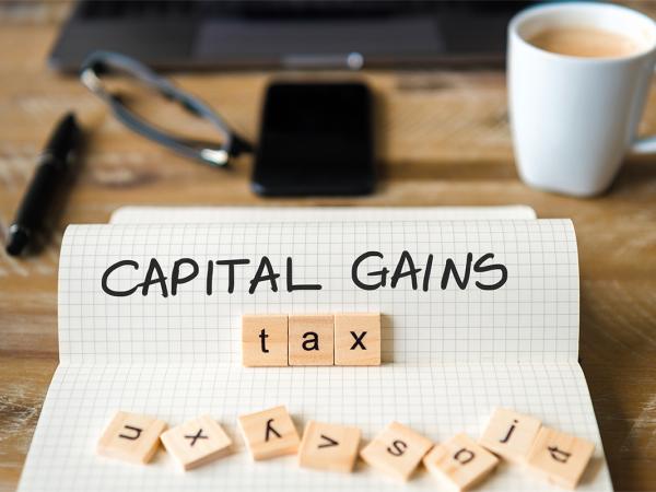 A pad of squared paper with the words 'CAPITAL GAINS' written on it in black ink, wooden scrabble tiles are scattered over the bottom of the paper with the word 'TAX' arranged to spell out 'CAPITAL GAINS TAX' 