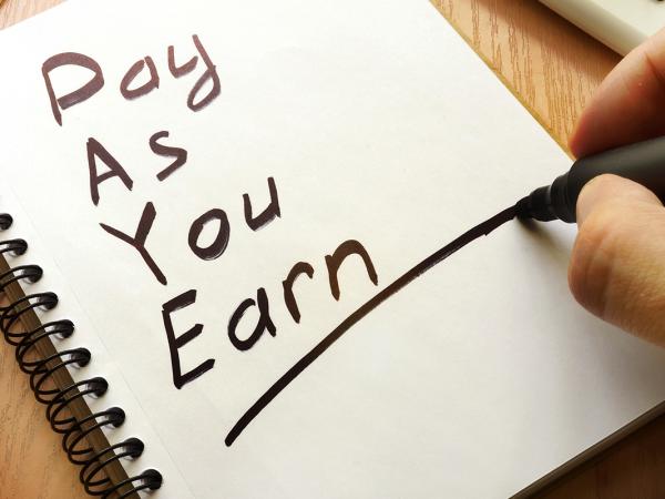 A person writing on a pad of white paper, the words 'PAY AS YOU EARN' are written in black handwriting and underlined.