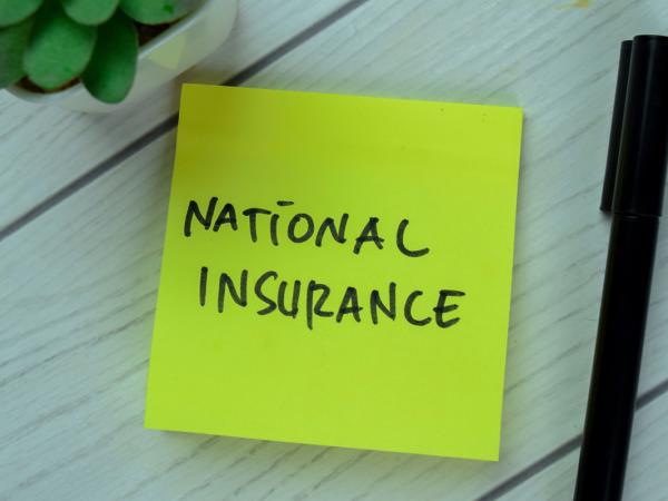 a yellow post it note with the words 'NATIONAL INSURANCE' written on it in black ink, a black pen sits next to this and also a small plant. 