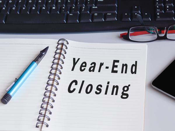 A pad of paper on a desk with the words 'YEAR-END CLOSING' typed on it. also on the deak a pen, a phone, a pair of glasses, a mouse mat and mouse and a keypad can be seen.