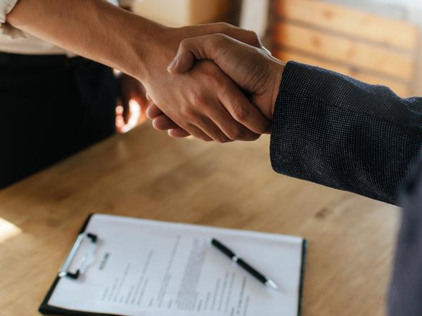 2 people shaking hands over a desk, on the desk some paperwork can be seen.