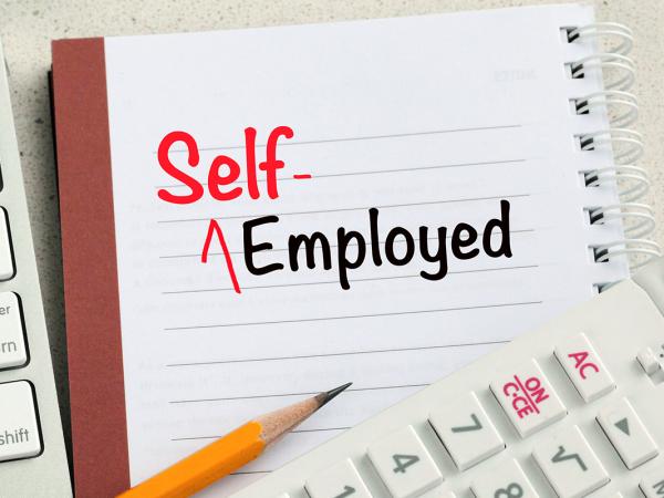 A keyboard, calculator, pencil and a pad of paper with the words 'SELF-EMPLOYED' written on it in black and red ink. 