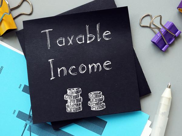 a piece of black paper on a desk surrounded by various stationary and graphs, the paper reads 'TAXABLE INCOME' in white writing along with an image of 2 stacks of coins. 