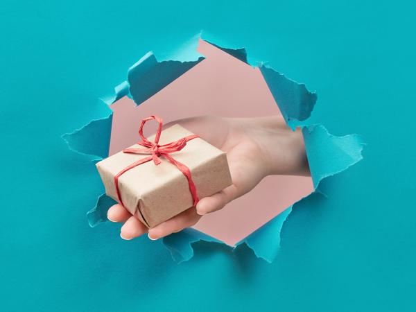 a blue paper wall with a hole in the middle, a persons hand appears through the hole holding out a gift box wrapped in brown paper with red string. 