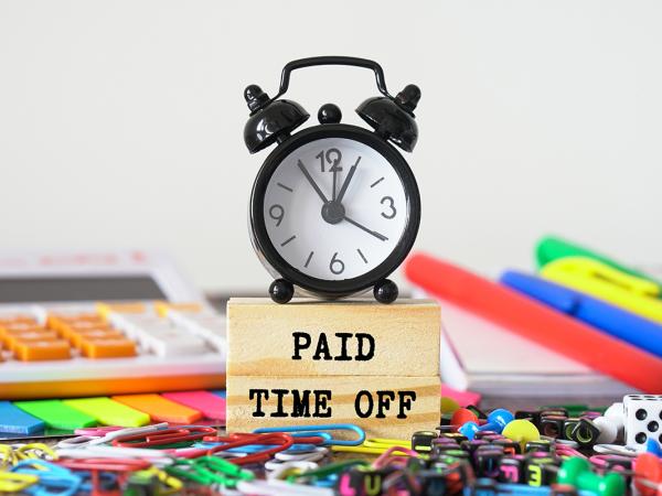 a desk scattered in various stationary, in the centre of the desk is a wooden block with the words 'PAID TIME OFF' and a clock sat on top.