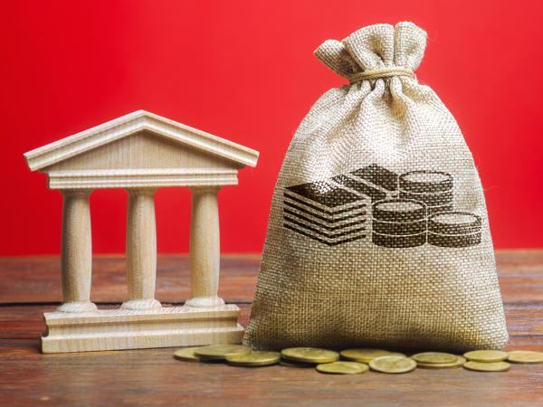 a red background and wooden table, a brown sack with an image of money drawn on the front sits next to a wooden figure of a state building, in front of this a scattering of coins can be seen.