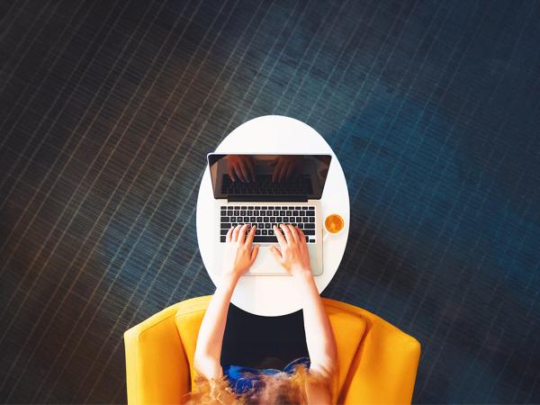 A birds eye view photo of a person sat on a yellow chair typing on a laptop that sits on top of a small white oval table, a dark checked pattern covers the carpet. 