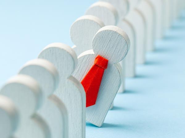 a line of wooden people, one wearing a little red tie is poking out from the line. 