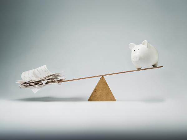 a man made see saw against a neutural coloured background. on one side is a piggy bank, the other side has papers. Image shows the papers are heavier that the piggy bank. 