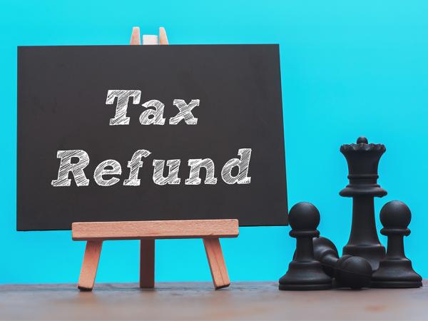 A wooden easel with a chalk board propped on it, the words 'TAX REFUND' are written in white chalk. next to this are some black chess pieces. Behind is a light blue background.