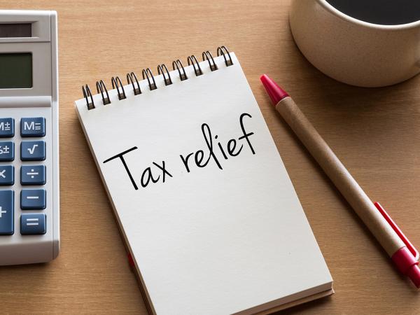 a desk with a calculator, a pen and a cup of coffee, in the centre is a notepad with the words 'TAX RELIEF' written on it. 