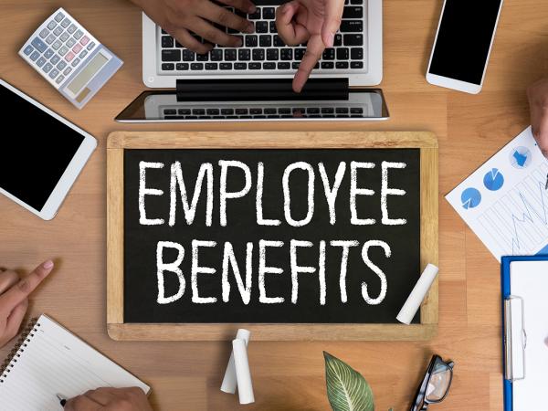 3 people sat around a desk, on the desk is various paperwork, stationary and electronic devices, in the centre of the desk is a chalkboard with the words 'EMPLOYEE BENEFITS' written on it in white chalk. 