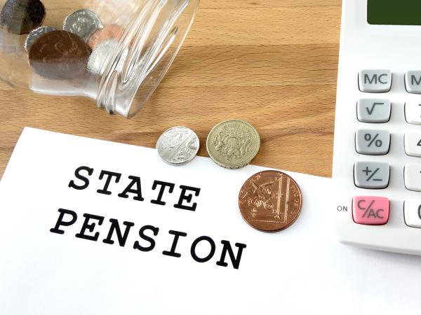a sheet of white paper with the words 'STATE PENSION' typed on it, next to this is a glass jar of coins tipped onto its side, a few of the coins have fallen out onto the paper, a person is typing on a calculator at the right hand side. 