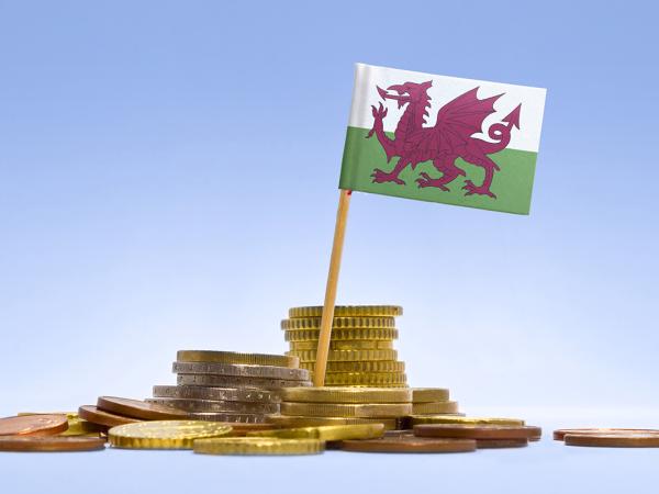 a pile of coins against a blue background, a Welsh flag is stood amongst the pile of coins. 