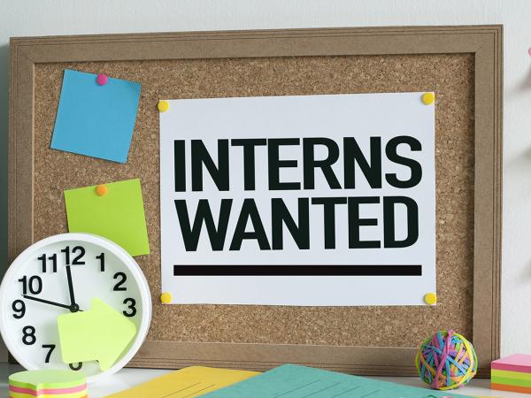a message board with the words 'INTERNS WANTED' pinned to it,  across the desk various stationary can be seen and a clock. 