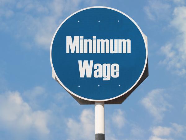 a blue sky with clouds and a blue circular road sign with the words 'MINIMUM WAGE' written on it in white text. 