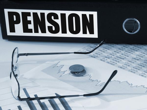 a thick black folder with the word 'PENSION' written on the spine sat on a desk with a pair of glasses, paperwork, a calculator and a jar of coins.