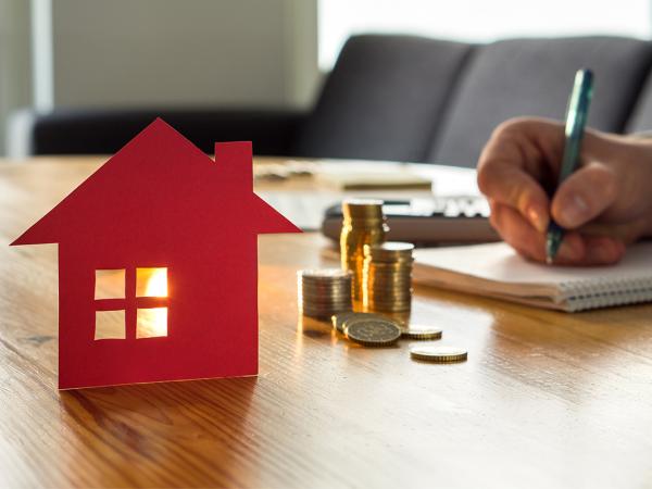 A person sat at a desk writing, also on the desk is a small red house and a stack of coins. 