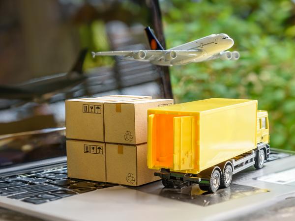 A laptop with tiny cardboard boxes and a toy haulage truck sat on the keyboard, a toy aeroplane is flying over the top of those.