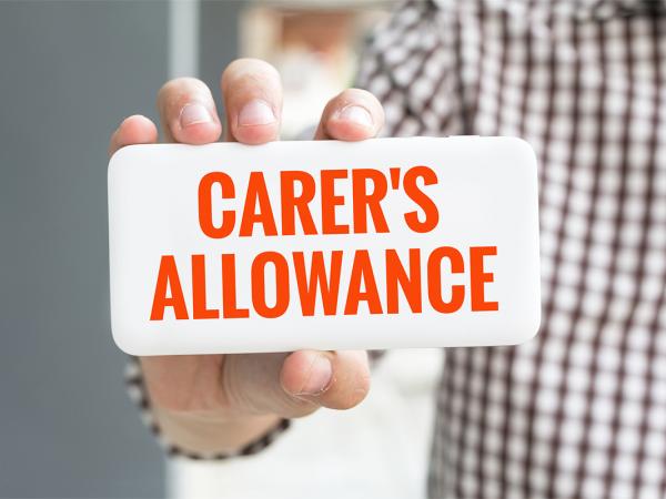 a person holding up a white card that says 'CARER'S ALLOWANCE' in orange text