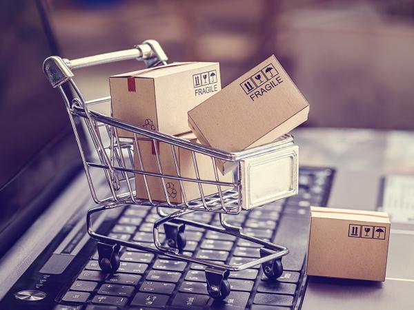 an open laptop with a tiny shopping trolley sat on the keyboard, inside the trolley are tiny brown parcels. 