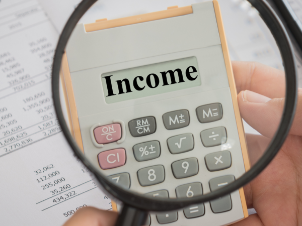 a person holding a magnifying glass up to a calculator showing the word 'INCOME' on the screen, behind this paperwork can be seen.
