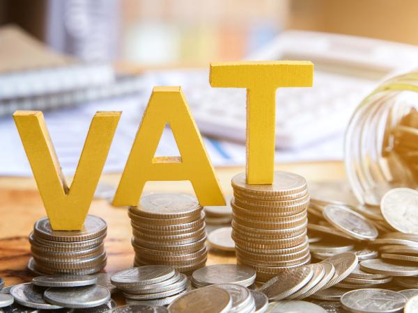 A desk scattered with coins and notebooks, a glass jar of coins is tipped on its side, three piles of coins can be seen in the centre each with a yellow letter on top. Together this spells 'VAT'.