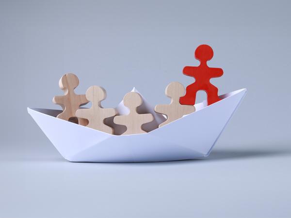 a paper boat with small wooden people inside, the person at the front of the boat is coloured red. 