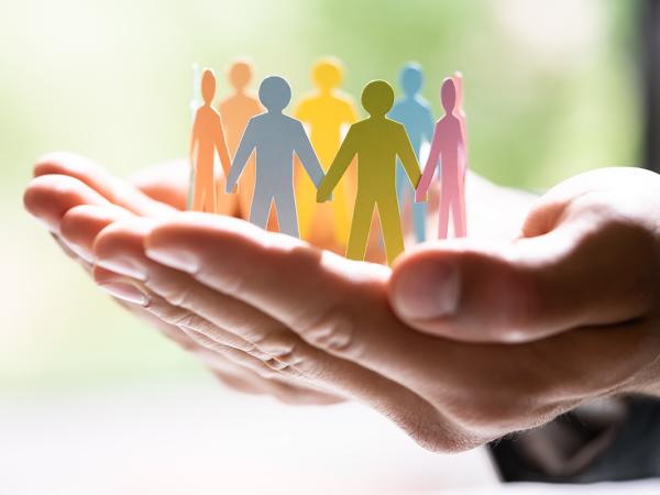 A person holding a paper chain of pastel coloured people holding hands stood in a circle. people are coloured in Pink, Green, Blue, Orange, Yellow. 