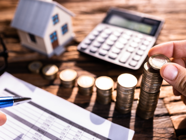 A person at a desk holding a pen with paperwork, stacks of coins, a small toy house and a calculator.