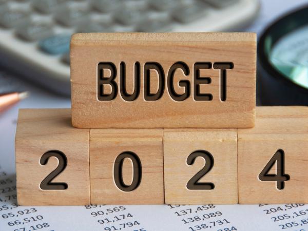 stacked wooden blocks, each block has engraved writing in black text on the side that together reads 'BUDGET 2024' background briefly shows a calculator, magnifying glass, a pen and paperwork. 