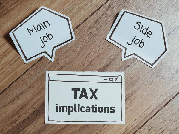 3 pieces of paper on a wooden desktop, one says 'TAX IMPLICATIONS', the other two are shaped like speech bubbles one saying 'MAIN JOB'. the other 'SIDE JOB' 