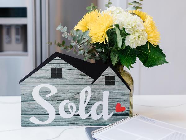 a bunch of flowers and a model house with the word 'SOLD' on the front sat on a kitchen countertop.