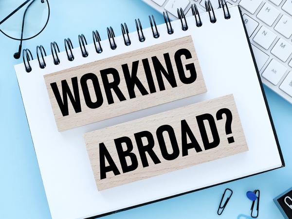 Two wooden blocks with the words 'WORKING ABROAD' showing on them in black text  sat on top of a notebook on a persons desk. Scattered around the notebook various stationary can be seen and a computer keyboard.