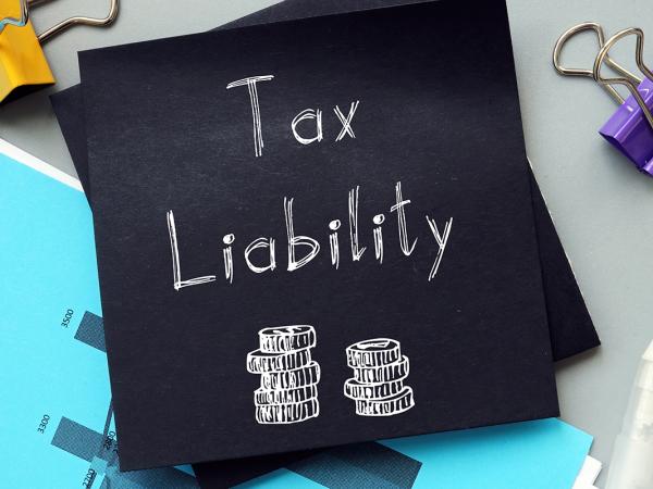 Black sheet of paper with the words 'TAX LIABILITY' written in white with an illustration of 2 piles of coins, various stationary surrounds the piece of black paper. 