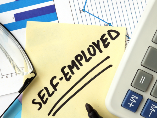 A desk of stationary along with a post-it note that reads 'SELF-EMPLOYED'.
