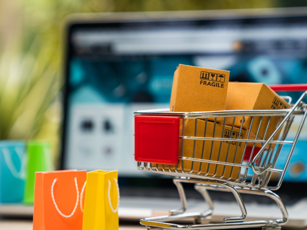 various different coloured shopping bags, a shopping trolley full of cardboard boxes and a laptop screen in the background. 