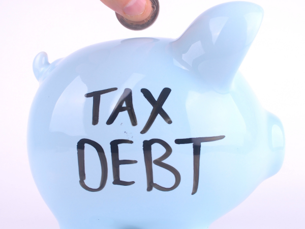 A person putting coins into a light coloured piggy bank with the words 'TAX DEBT' written in black ink. 