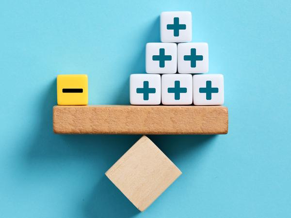A wooden see saw  perfectly balanced against a blue wall, on one side of the shelf is a yellow block with a minus sign on it, on the other side there are 9 white blocks with plus signs on each block.